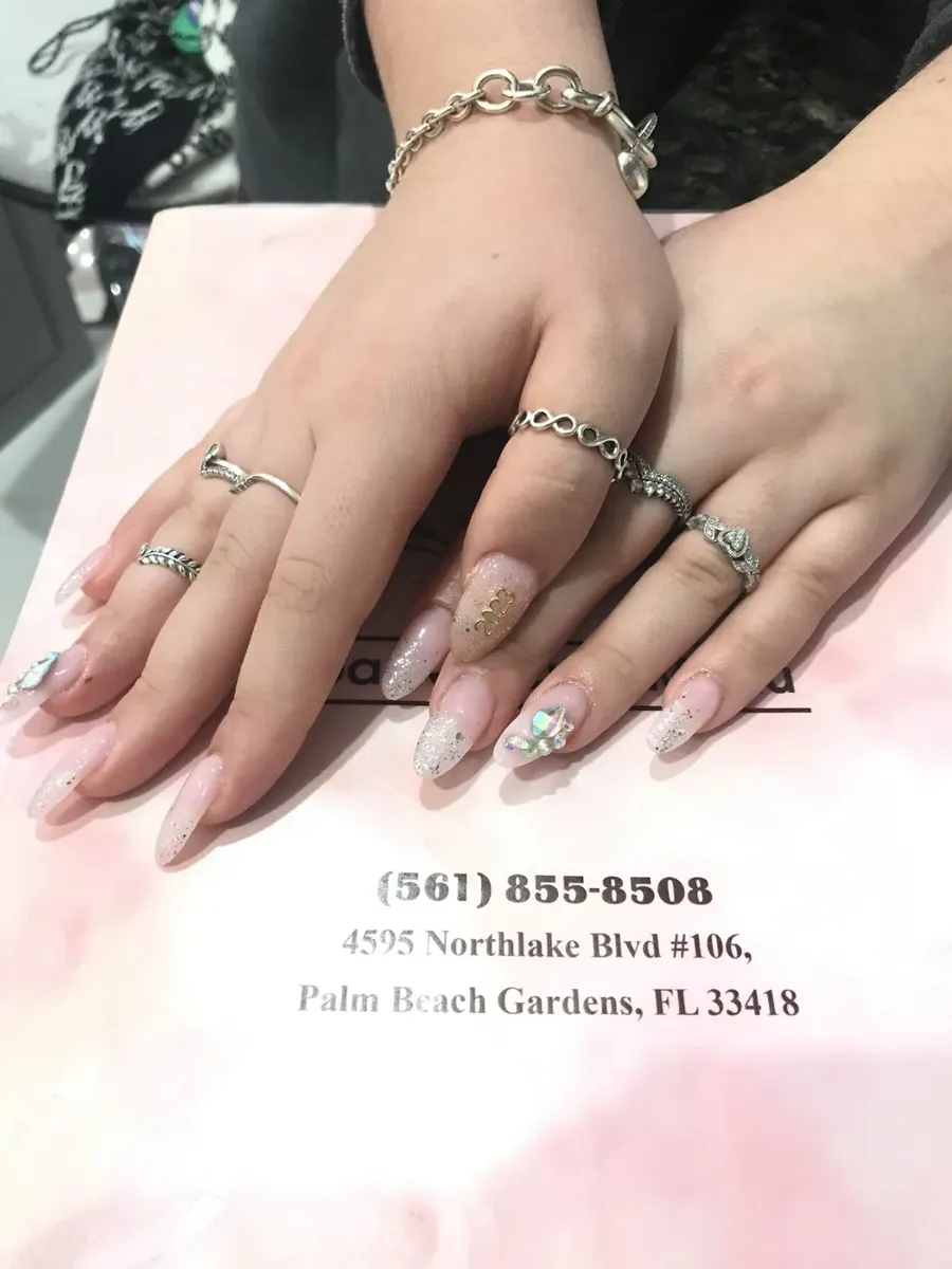 Hands with long almond nails in pink glitter fading to clear, with crystal accents, resting on the salon's pink card